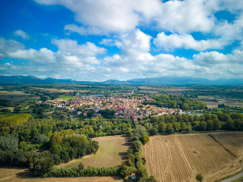 Aerial view of the historic town of Peralada surrounded by lush green forests, agricultural fields, and rolling hills under a bright blue sky in Peralada, Catalonia, Spain.