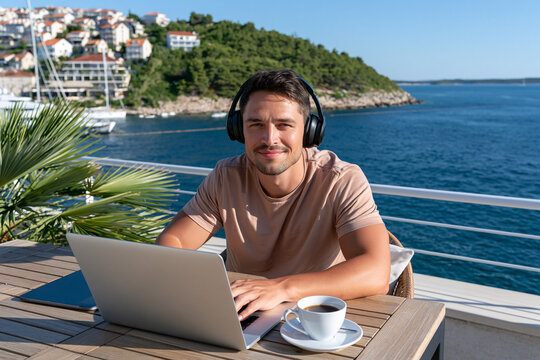 Man smiles while working on a laptop outdoors. He wears headphones and sips coffee. Bright blue water and green hills frame the scene. He sits at a wooden table near a railing