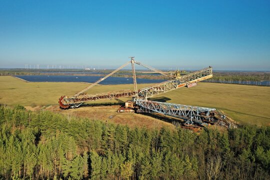 Aerial view of a massive bucket-wheel excavator standing in the Lausitz region, surrounded by open-pit mining fields, forests, and a clear blue sky in Brandenburg, Germany.