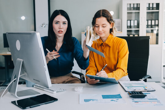 Two businesswomen discussing work using a laptop in a modern office lounge area, showing teamwork and communication