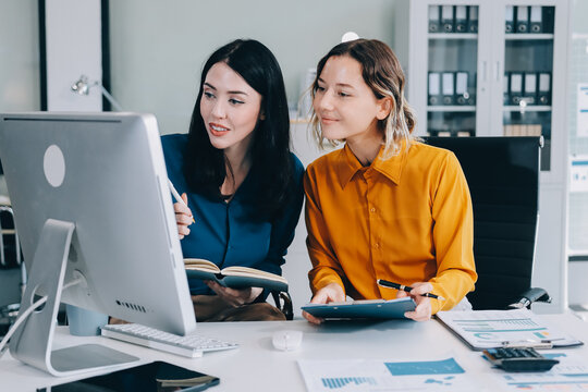 Two businesswomen discussing work using a laptop in a modern office lounge area, showing teamwork and communication