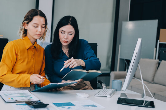 Two businesswomen discussing work using a laptop in a modern office lounge area, showing teamwork and communication