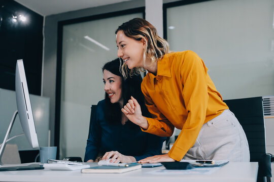 Two businesswomen discussing work using a laptop in a modern office lounge area, showing teamwork and communication