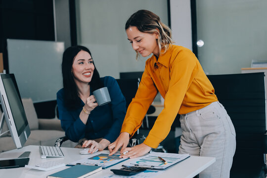 Two businesswomen discussing work using a laptop in a modern office lounge area, showing teamwork and communication