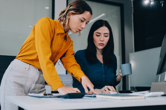Two businesswomen discussing work using a laptop in a modern office lounge area, showing teamwork and communication