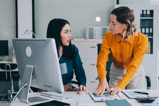 Two businesswomen discussing work using a laptop in a modern office lounge area, showing teamwork and communication