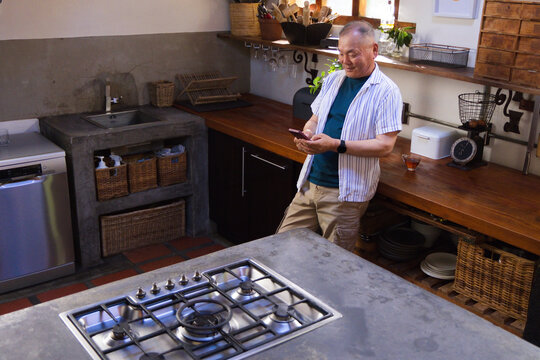 Senior Asian male leaning on countertop by window, checking smartphone near cooktop, copy space