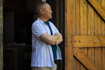 Fototapeta premium Senior Asian man standing in shed doorway by wood door, crossing arms, wearing striped shirt watch