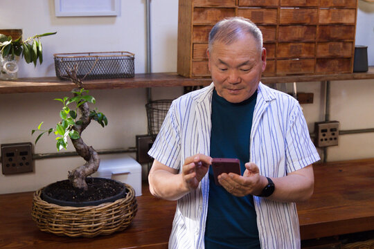 Senior Asian man standing in workshop at wooden counter, holding smartphone near bonsai basket