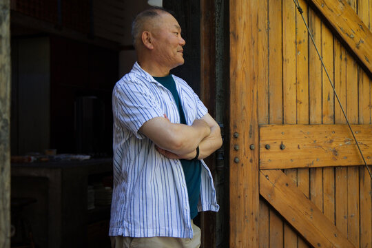 Senior Asian man standing in shed doorway by wood door, crossing arms, wearing striped shirt watch
