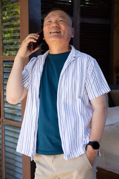Senior Asian man standing in porch by louvered shutters, holding smartphone to right ear, smiling
