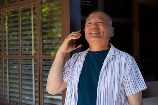 Senior Asian man holding phone to right ear, smiling, looking up on porch by louvers, striped-shirt