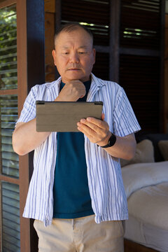 Senior Asian man standing and holding tablet at chest on covered porch near louvered shutters