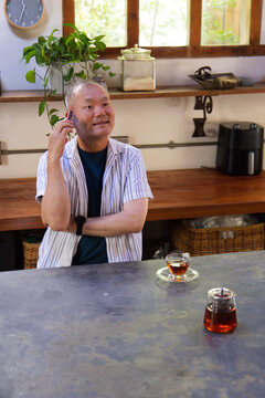 Senior Asian man sitting at wooden countertop in home kitchen, holding smartphone and tea carafe