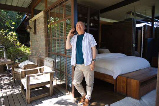 Senior Asian man standing on wooden veranda holding smartphone to ear, wristwatch, low bed visible