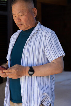 Senior Asian man standing in dim room, looking at smartphone, wearing striped shirt and watch
