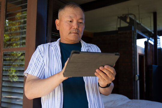 Senior Asian man standing on veranda at home holding tablet in folio wearing wristwatch