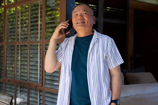 Senior Asian man standing on porch near louvered window, holding smartphone, wearing wristwatch