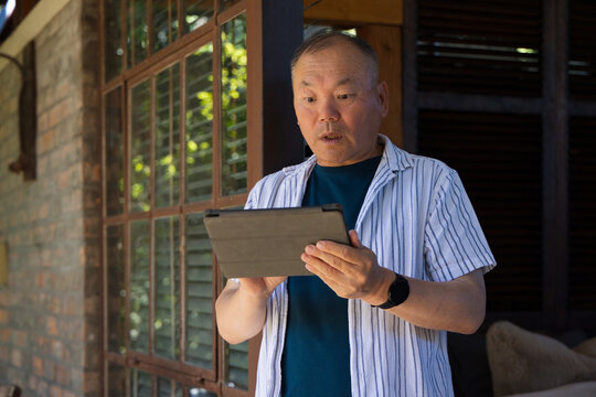 Senior Asian man standing on porch holding tablet and wearing striped shirt near glass door