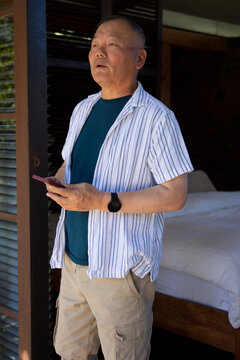 Senior Asian male standing at bedroom threshold holding smartphone with black smartwatch visible
