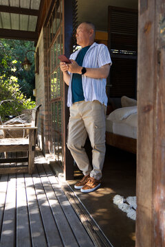 Senior Asian man standing in cabin doorway holding smartphone and smartwatch on wooden deck