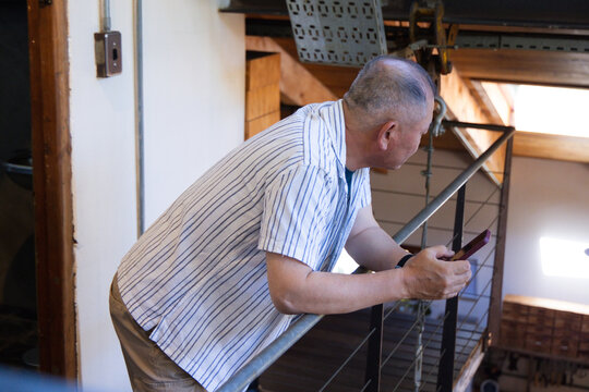 Senior Asian man leaning on metal rail in loft holding smartphone wearing striped shirt and watch