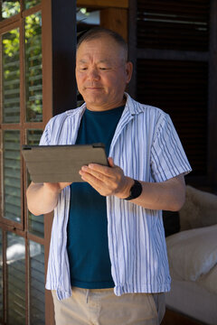 Senior Asian man standing by wooden door holding tablet, wearing striped shirt and smartwatch