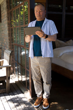 Senior Asian man standing in doorway holding tablet, wearing striped shirt and smartwatch