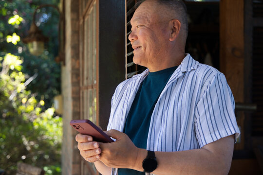 Senior Asian man standing in doorway wearing teal tee and black watch, holding pink phone, gazing