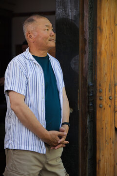 Senior Asian man standing in doorway, clasping hands, looking right near wooden door panel