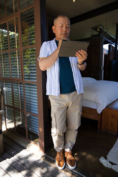 Senior Asian man standing in doorway holding tablet, looking at screen on wooden deck