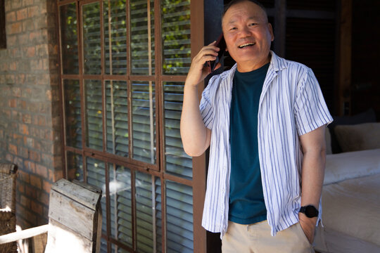 Senior Asian man standing in doorway on porch near wooden shutters holding smartphone, copy space