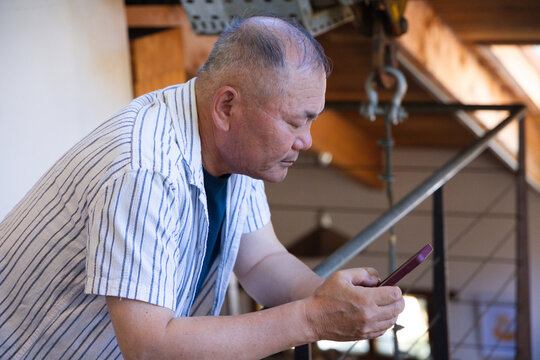 Senior Asian man leaning on metal railing, wearing striped shirt, holding maroon smartphone in loft