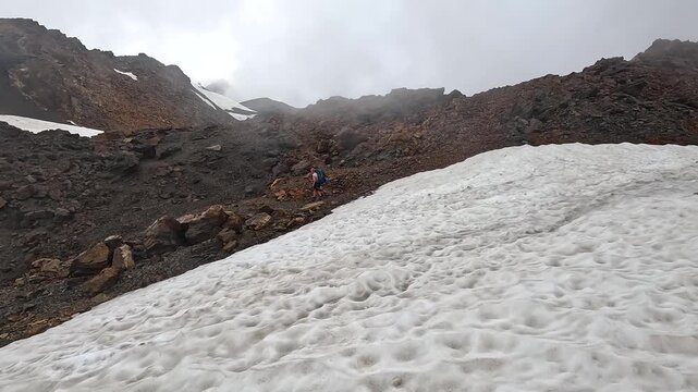 Hiker walks rocky slope, wide view beside a remaining snowfield in Svaneti, Georgia, documenting alpine route navigation, seasonal trail conditions and trekking tourism across rugged Caucasus terrain