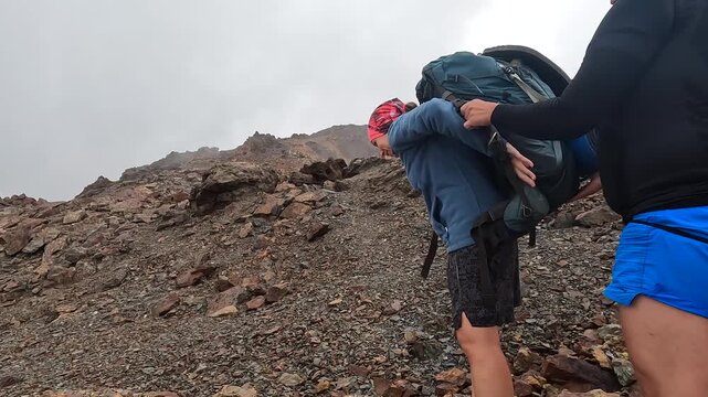 Male hiker assists partner, side view, with lifting a heavy backpack on a rocky slope in Svaneti, Georgia, showing teamwork, equipment handling and mutual support during expedition preparation