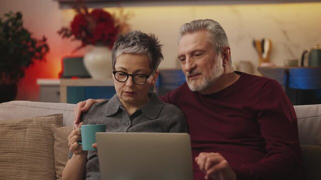 Positive couple in their 50s shop on laptop at home, booking hotel tickets