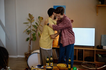 Naklejka premium Diverse young adult male football fans hugging in living room while celebrating game victory, representing friendship and sports entertainment, ideal for lifestyle marketing campaigns