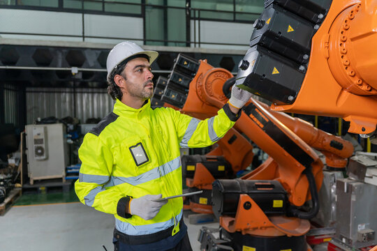 Technician wearing hardhat and safety vest holding tablet to inspect robotic arm in factory.
