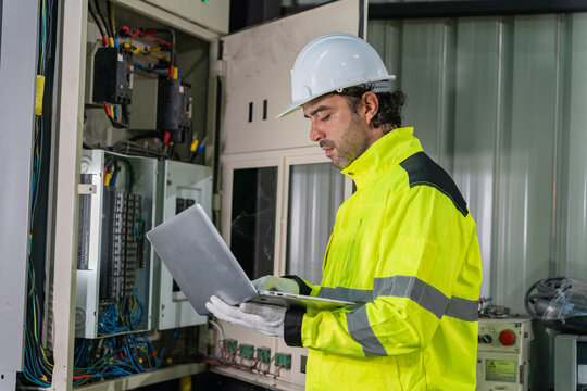 Technician wearing hardhat using laptop computer to inspect electrical control cabinet in factory.