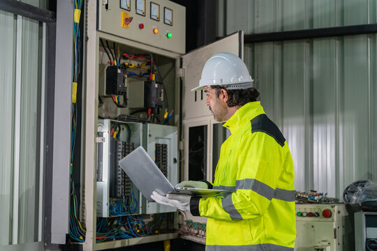 Technician wearing hardhat using laptop computer to inspect electrical control cabinet in factory.