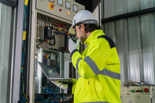 Technician wearing hardhat using laptop computer to inspect electrical control cabinet in factory.