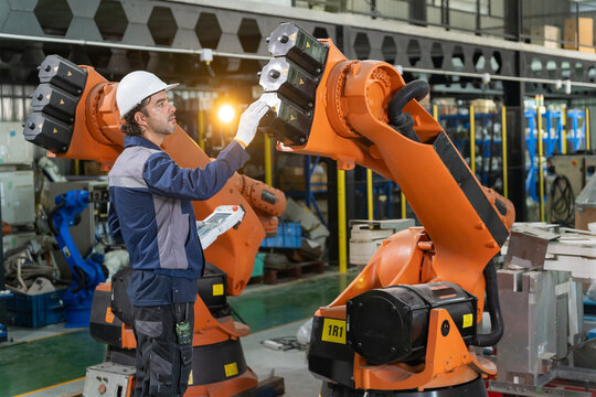 Technician wearing hardhat using digital control pendant to inspect and program robotic arm in factory.