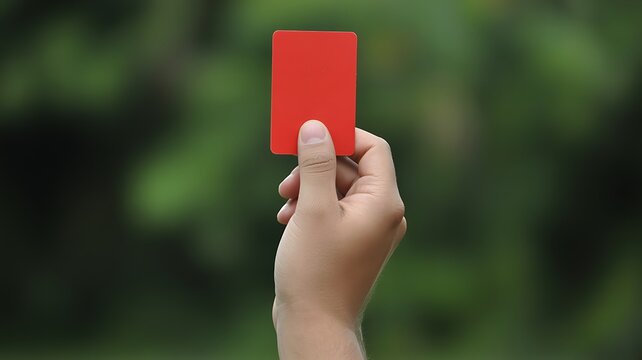 A close-up photograph of a hand holding a red penalty card signifying a sports foul or disciplinary action in an outdoor setting