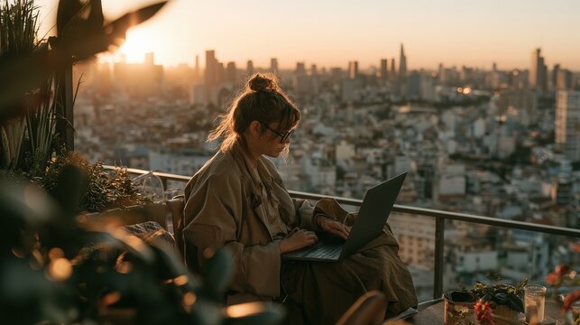 Woman with a bun works on her laptop from a high-rise rooftop terrace, enjoying the panoramic city skyline view under the golden light of dusk.