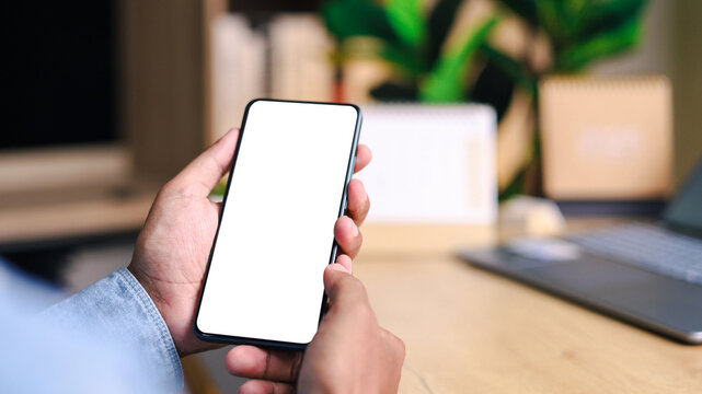 Hand holding a blank smartphone with a clean white screen, laptop and green plant in a cozy workspace, ideal for mobile technology, business, app, and digital lifestyle concepts.
