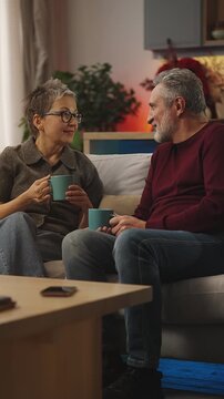 Happy couple in their 50s talk while drinking tea, relaxing together on sofa