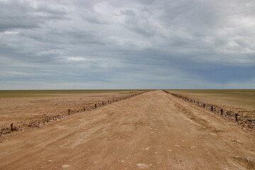 Eine gerade, unbefestigte Piste führt durch eine weite flache Landschaft mit niedrigem Horizont und dicht bewölktem Himmel. © Jane Be. The Picture