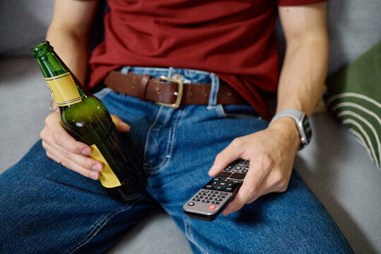 Cropped shot of young adult Caucasian male football fan holding beer bottle and TV remote control while sitting on sofa. Perfect for sports broadcasting and entertainment marketing