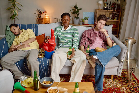 Diverse young adult male football fans sitting on sofa looking disappointed after game loss. Image suitable for sports marketing campaigns and social media content