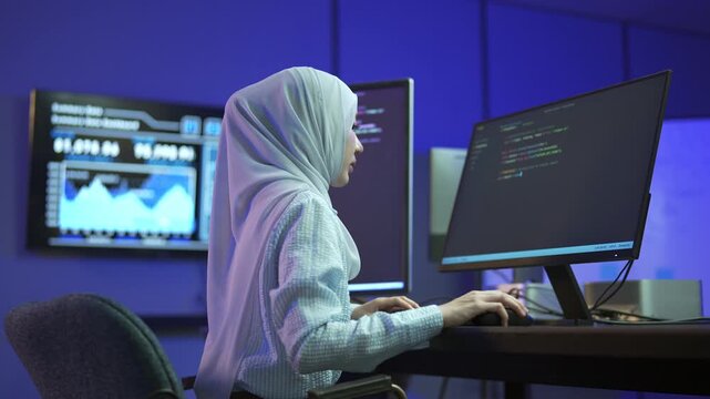 A stressed Muslim woman programmer in hijab has headache while writing program code on multiple desktop screens in a modern office.